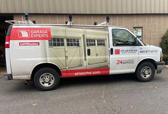 White and red service van for Atlas-Coles Door Systems, with Garaga and LiftMaster logos, promoting 24-hour emergency garage door service. It displays a phone number and website, parked outside a brick commercial building.